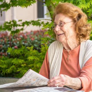An elderly woman happily reading a newspaper in a garden setting.