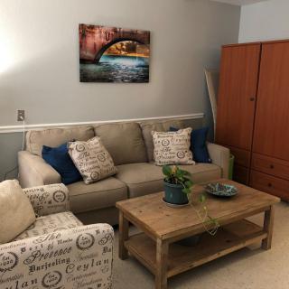 A cozy living room with a sofa, armchair, and coffee table, adorned with cushions and plants. An artwork hangs on the wall.