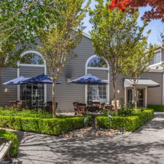 A charming outdoor patio area with tables shaded by umbrellas, surrounded by neatly trimmed bushes and trees.