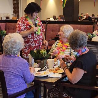 A group of seniors enjoying a meal together, adorned with leis, engaging in lively conversation.