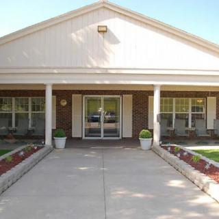 The image shows the entrance of a building with a covered porch, symmetrical windows, and neatly landscaped surroundings.