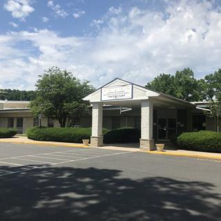 Entrance of a building surrounded by trees and a parking lot under a partly cloudy sky.