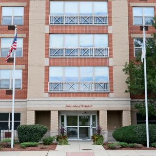 A modern multi-story building with flags at the entrance, surrounded by trees and shrubs.