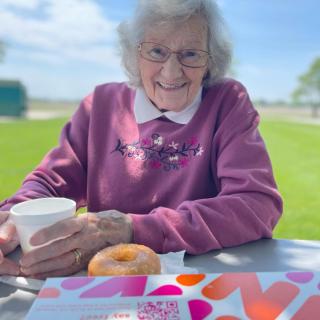 An elderly woman enjoys coffee and a donut while sitting outside on a sunny day.