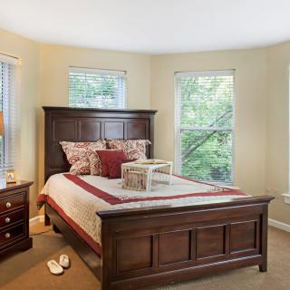 A cozy bedroom featuring a wooden bed with decorative pillows, illuminated by natural light from the windows.
