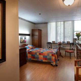 A cozy bedroom with a single bed and colorful quilt, featuring wooden furniture and a large window with blinds.