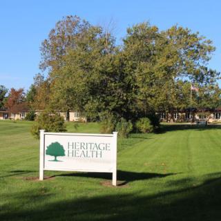 A sign for Heritage Health stands on a well-maintained lawn with trees and a building in the background.