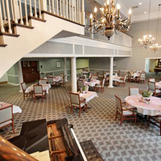 An elegant dining area with chandeliers, carpeted floors, and a piano, featuring tables and chairs arranged for guests.