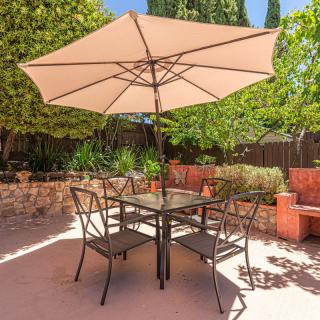 A sunny backyard patio with a dining set featuring an umbrella, table, and chairs surrounded by greenery.