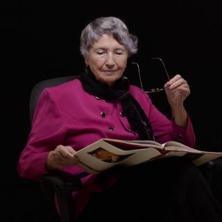 An elderly woman in a pink blazer is seated and reading a book while holding her glasses.