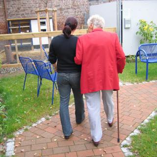 A caregiver assisting an elderly person while walking in a garden.