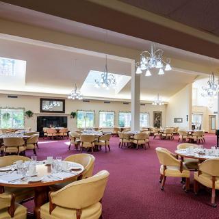 A spacious and well-lit dining area with multiple tables and chairs, featuring elegant chandeliers and a rich carpeted floor.