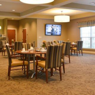 A spacious dining area with neatly arranged tables and chairs, illuminated by overhead lights, featuring a television on the wall.