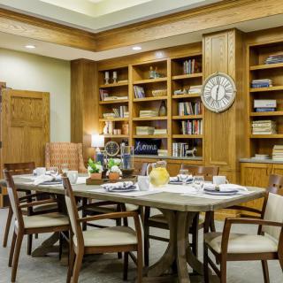 A dining area with a wooden table set for a meal, surrounded by bookshelves and a clock on the wall.