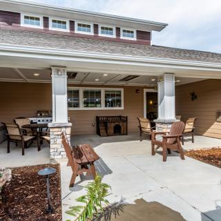 A well-furnished patio with outdoor seating and tables, featuring columns and surrounding landscaping.