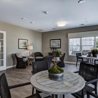 A cozy seating area with multiple tables and chairs, decorated with plants and art, featuring large windows for natural lighting.