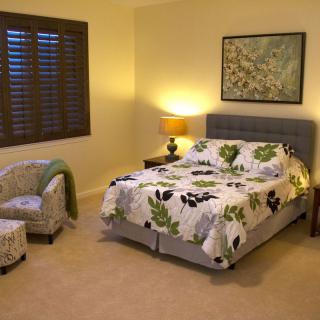 A cozy bedroom featuring a bed with floral bedding, a reading chair, side table with a lamp, and artwork on the wall.