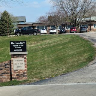 A view of a facility with a sign indicating its name, surrounded by grass and trees, with several parked vehicles.