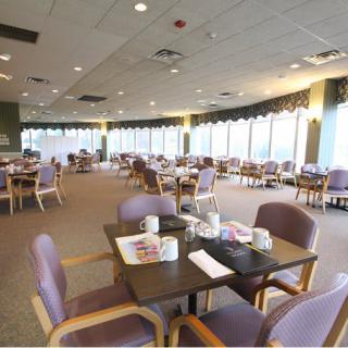 An empty dining hall with neatly arranged tables and chairs, featuring large windows.