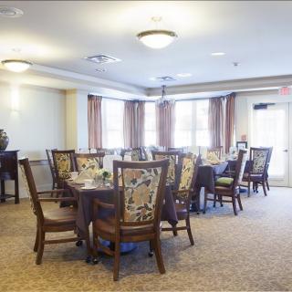 A well-lit dining room with multiple tables and chairs, featuring elegant curtains and a carpeted floor.