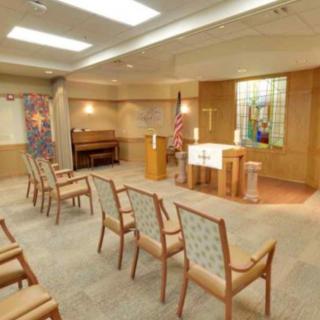 A small chapel interior with seating arranged around a central altar and a stained glass window in the background.