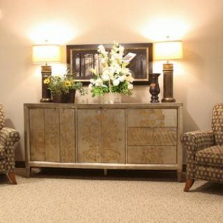 A warmly lit living room with a sideboard, lamps, floral arrangement, and patterned chairs.
