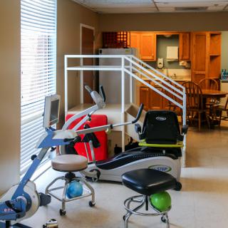 A well-equipped rehabilitation space featuring exercise machines and a wheelchair ramp, with a view of a kitchen in the background.