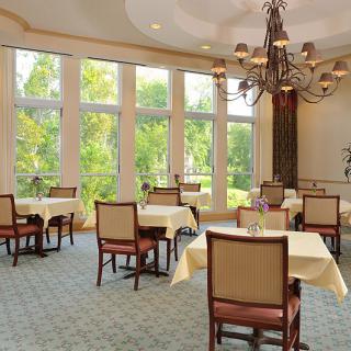 A well-lit dining room with several tables and chairs, featuring large windows and an elegant chandelier.