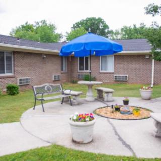 A cozy outdoor patio area featuring a bench, stone table with chairs, and a blue umbrella surrounded by potted plants.