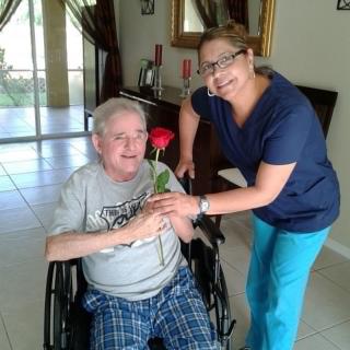 A caregiver giving a flower to an elderly man in a wheelchair, both smiling warmly inside a cozy home.