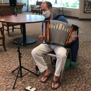 A musician wearing a mask plays the accordion indoors while sitting on a chair.