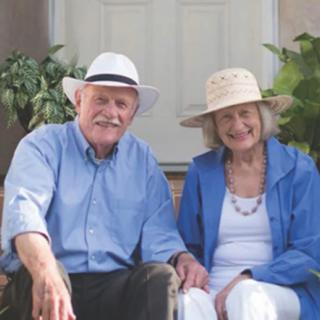 An elderly couple sits happily outdoors, both wearing hats.