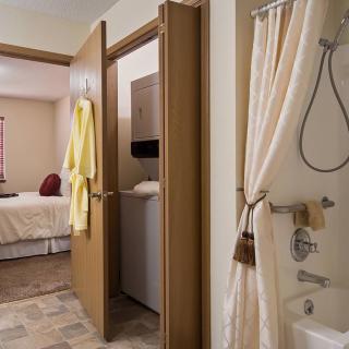 A view from a bathroom showing a shower with a curtain, towels hanging on doors, and an adjacent bedroom.