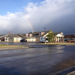 A rainbow appears over a building with a wet parking lot under a cloudy sky.