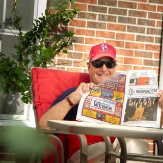 A person relaxing on a patio chair, reading a newspaper while wearing sunglasses and a cap.