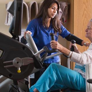 A therapist assists an elderly woman with exercise on fitness equipment.