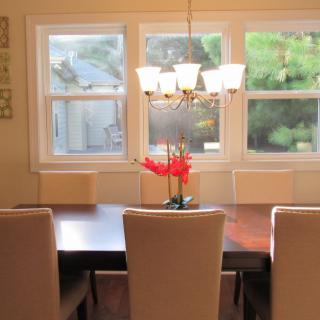A well-lit dining room featuring a large table with chairs and a chandelier, overlooking a scenic outdoor view through the windows.