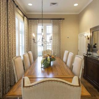 An elegant dining room featuring a long wooden table, surrounded by upholstered chairs and adorned with a central floral arrangement. Large windows with patterned curtains allow natural light to illuminate the space, complemented by a ceiling chandelier.