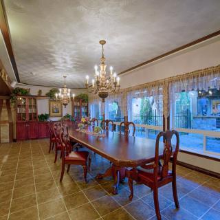 An elegant dining room with a long table, surrounded by chairs and illuminated by chandeliers, featuring large windows that allow natural light to stream in.
