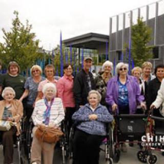A group of elderly individuals, some in wheelchairs, gathered outdoors in front of a building.