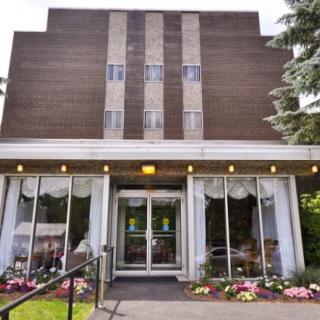 A building entrance with a brick facade, featuring large glass doors and surrounding landscaping with flowers and trees.