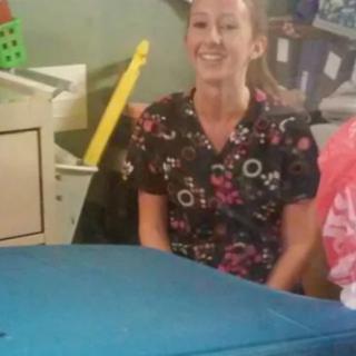 A woman wearing scrub attire smiles while sitting indoors in a work environment.