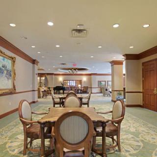 A spacious dining area with elegant furniture, featuring round tables and upholstered chairs, accented by a large painting and ceiling lights.