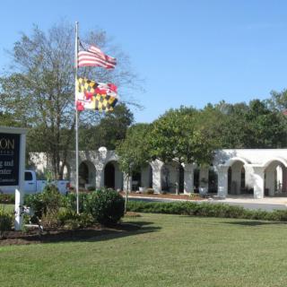 An outdoor scene featuring a building with arched windows, surrounded by trees and a manicured lawn, with American and Maryland flags flying near a sign.