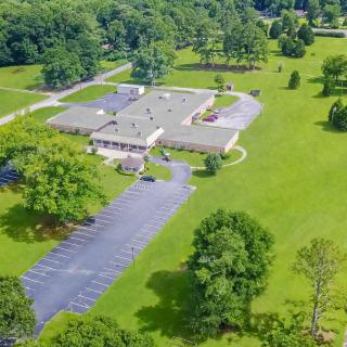 An aerial view of a building surrounded by greenery and trees, with an adjacent parking lot.