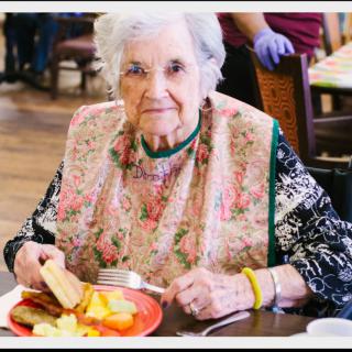 An elderly woman enjoys a meal at the dining table, wearing a floral bib.
