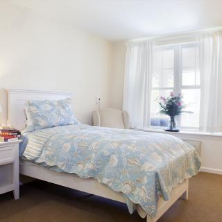 A bright and cozy bedroom featuring a single bed with floral bedding, a large window, and a lamp on the bedside table.