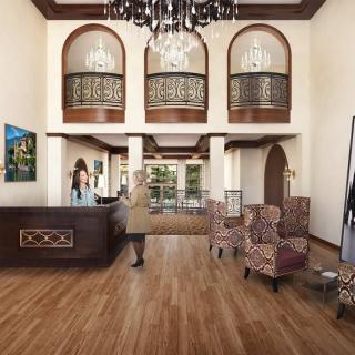 A spacious hotel lobby with elegant wooden flooring, featuring a reception desk, a seating area with patterned chairs, and decorative arches under a grand chandelier.