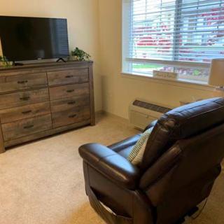 A cozy living room with an armchair facing a television on a dresser, next to a large window.