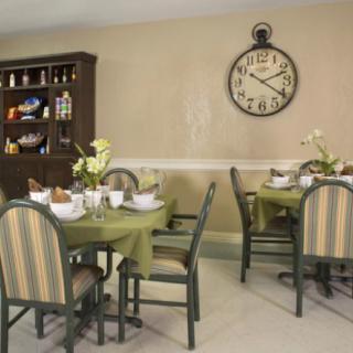 A dining room with a neatly set table, surrounded by chairs, a wall-mounted clock, and a cabinet in the background.